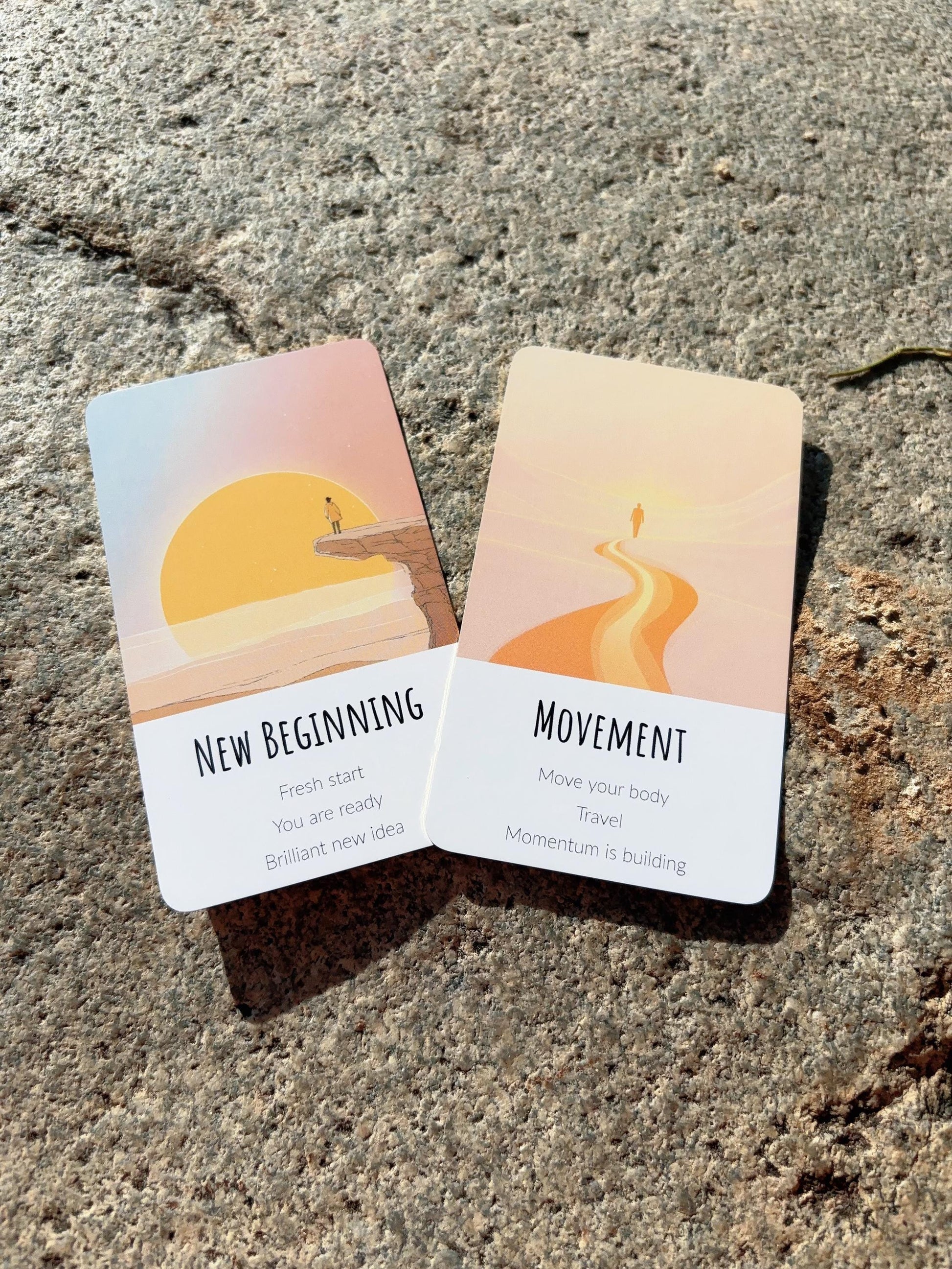 Abundance oracle cards with minimal illustrations and text messages. Two cards titled 'New Beginning' and 'Movement' on a textured surface.