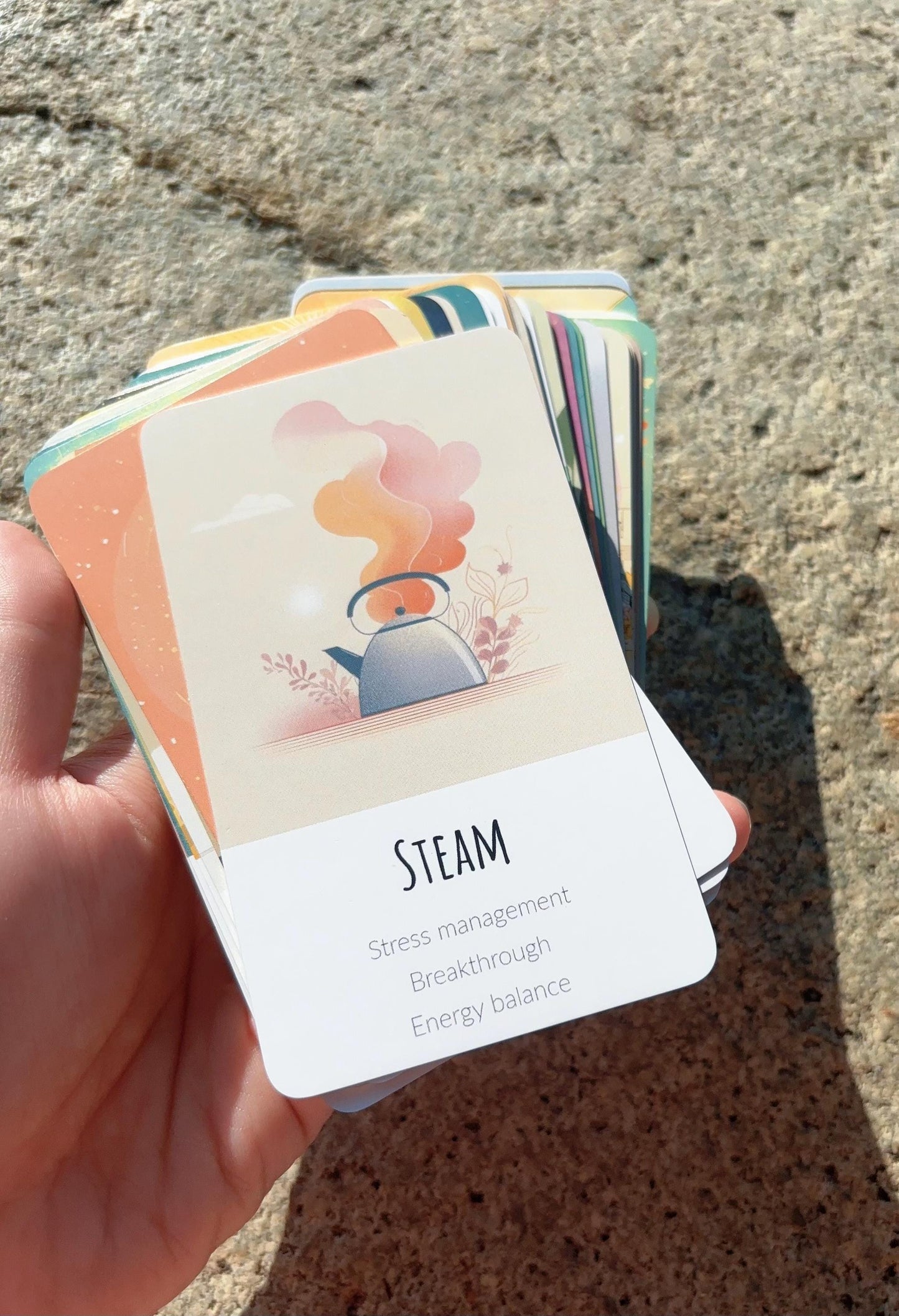 Abundance oracle cards for mindset and personal reflection displayed outdoors. Hand holding a card with 'STEAM' on it, featuring an illustration of a teapot and steam, against a concrete background.
