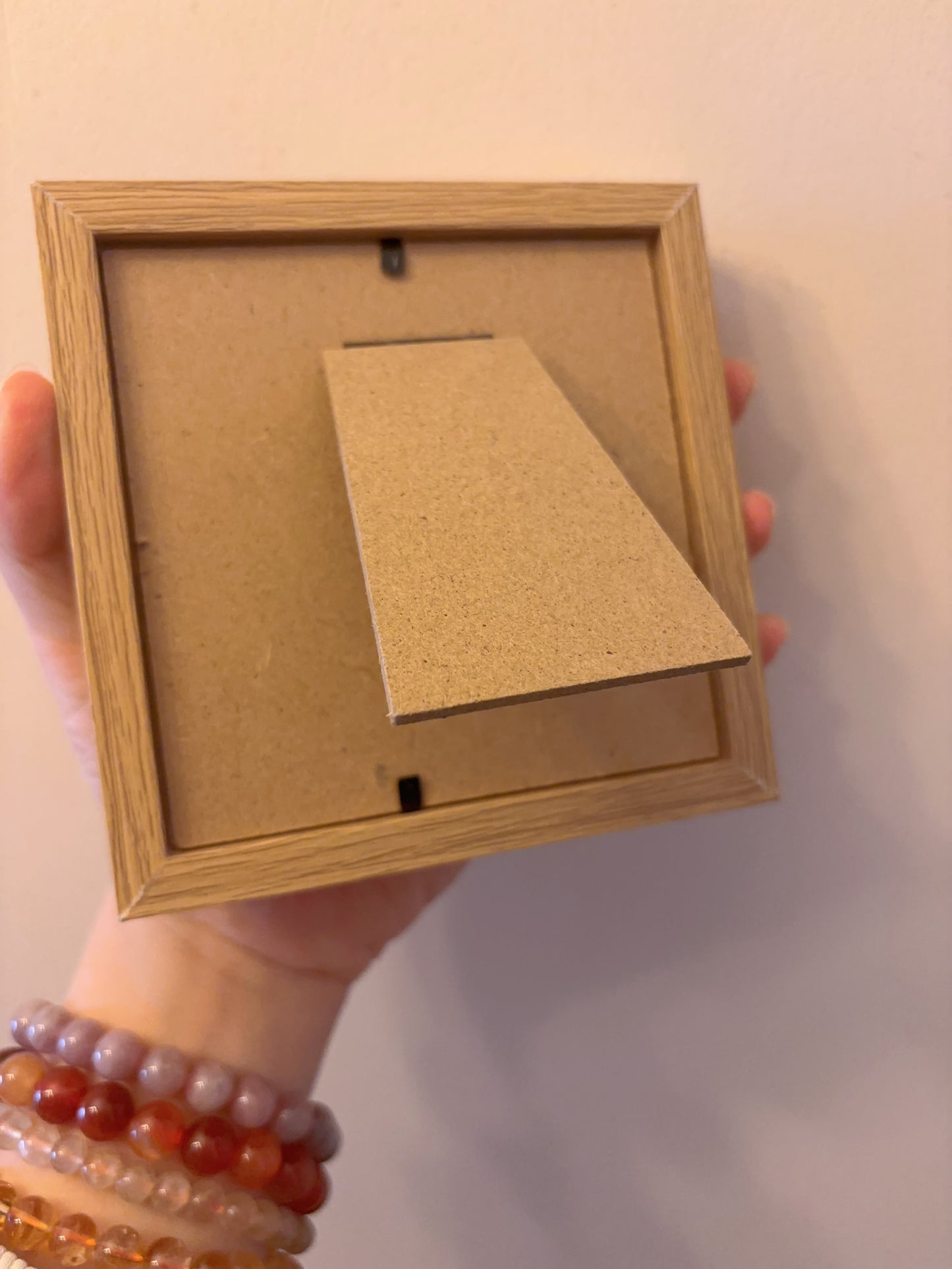 Hand holding a wooden picture frame with a plain cardboard insert and mini cosmos painting against a light background
